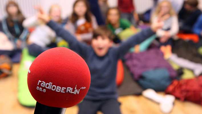 Reporter-Mikrofon von radioBERLIN, im Hintergrund sind Kinder auf Schlafmatten, ein Junge hebt beide Arme (Quelle: rbb/Matthias Bartsch)