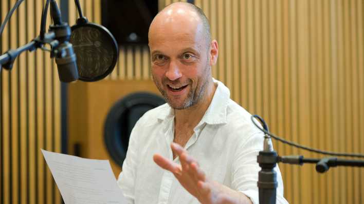 Schauspieler Christian Koerner im rbb-Studio beim Einsprechen hinter einem Mikrofon, weißes Hemd, Textblatt in der rechten Hand (Quelle: rbb/Oliver Ziebe)