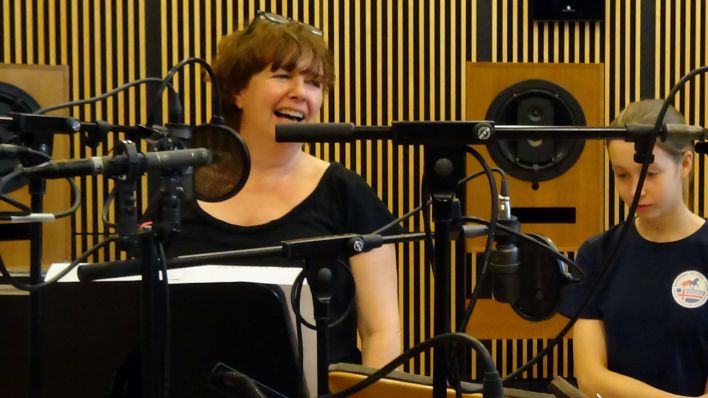 Frauke Poolmann, Erzählerin und Lehrerin, vor mehreren Mikrofonen im Studio; am Rande, rechts Lotta Stüber als Schülerin (Quelle: rbb/Robert Schoen)