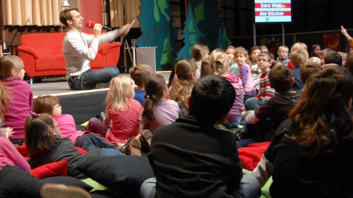 rbb-Moderator Marc Langebeck sitzt auf der Bühne mit dem Mikrofon, viele Kinder sitzen davor und hören zu © rbb/Redaktion OHRENBÄR