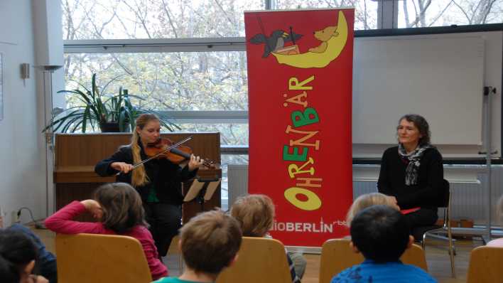 OHRENBÄR-Autorin Katrin Askan rechts und Musikerin Sarah Piorkowsky links neben dem OHRENBÄR-Banner, im Vordergrund kinder, die zuhören (Quelle: rbb/OHRENBÄR/Birgit Patzelt)