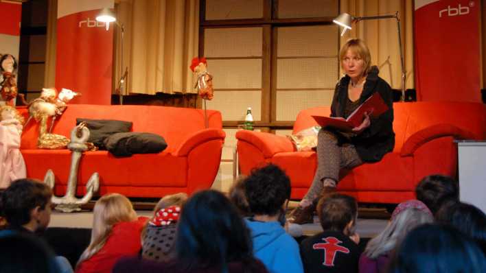 OHRENBÄR-Autorin Susanne Kornblum sitzt auf einer roten Couch und liest vor, im Vordergrund hören Kinder aufmerksam zu (Quelle: rbb/OHRENBÄR/Birgit Patzelt)