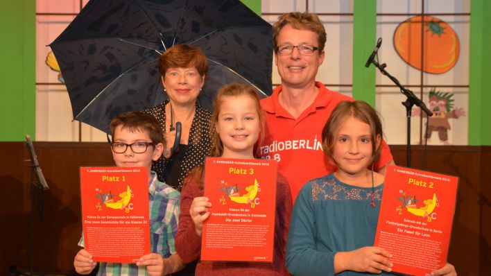 Zwei Schülerinnen und ein Schüler aus den Klassen der Presiträger des OHRENBÄR-Schreibwettbewerbs 2016, halten die Urkunden hoch, dahinter Schirmherrin Sabine Ludwig und Moderator Alexander Schurig (Quelle: rbb/Thomas Ernst)