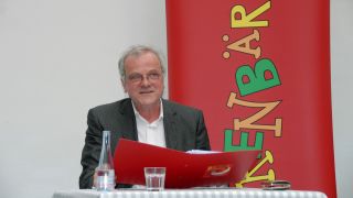 Schauspieler Bernhard Schütz am Tisch, er liest, im Hintergrund das OHRENBÄR-Banner (Quelle: rbb/OHRENBÄR/Birgit Patzel)
