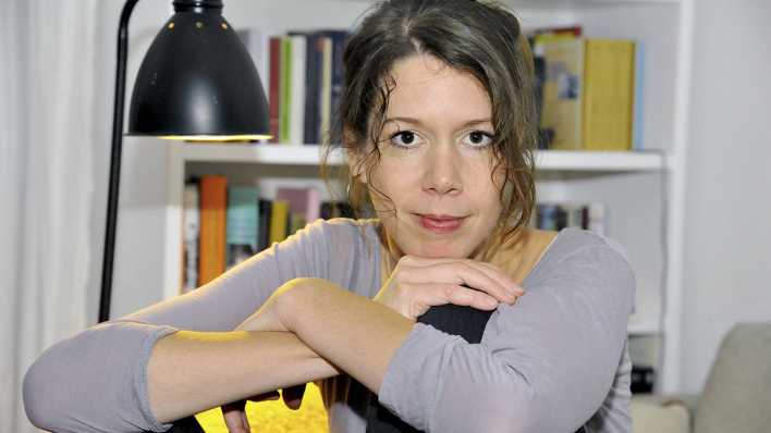 Porträt der Autorin Anna Jelena Schulte, vor einem Bücherregal, links daneben eine leuchtende Stehlampe (Quelle: privat)