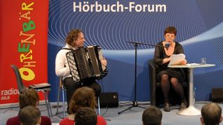 Autorin Susanne Friedmann liest, Musiker Uwe Steger am Akkordeon, im Hintergrund ein OHRENBÄR-Banner und der Schriftzug "Hörbuch-Forum" (Quelle: rbb/OHRENBÄR/Birgit Patzelt)