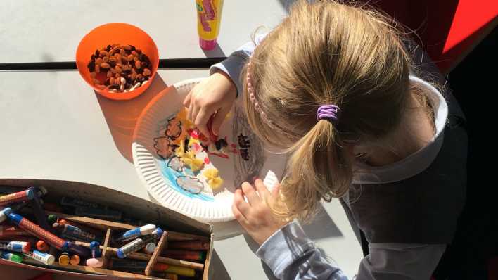 Ein Mädchen mit Zöpfen klebt Nüsse auf einen bunt bemalten Pappteller (Quelle: rbb/OHRENBÄR/Lisa Thieler)