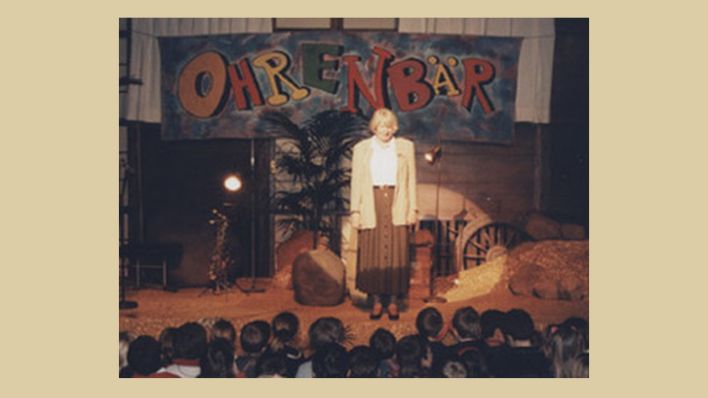 OHRENBÄR-Erfinderin Marianne Wagner auf der Bühne, im Hintergrund das erste OHRENBÄR-Wortlogo (Quelle: rbb/OHRENBÄR)